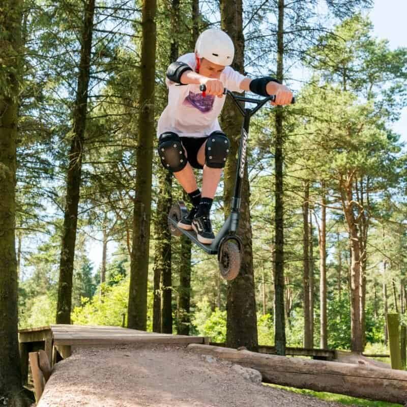 Tough guy on an e-scooter jumps from a wooden ramp in wooded area.