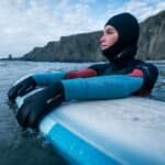 Entspannter Surfer im Neoprenanzug liegt auf dem Surfbrett am Meer mit felsigen Klippen im Hintergrund.