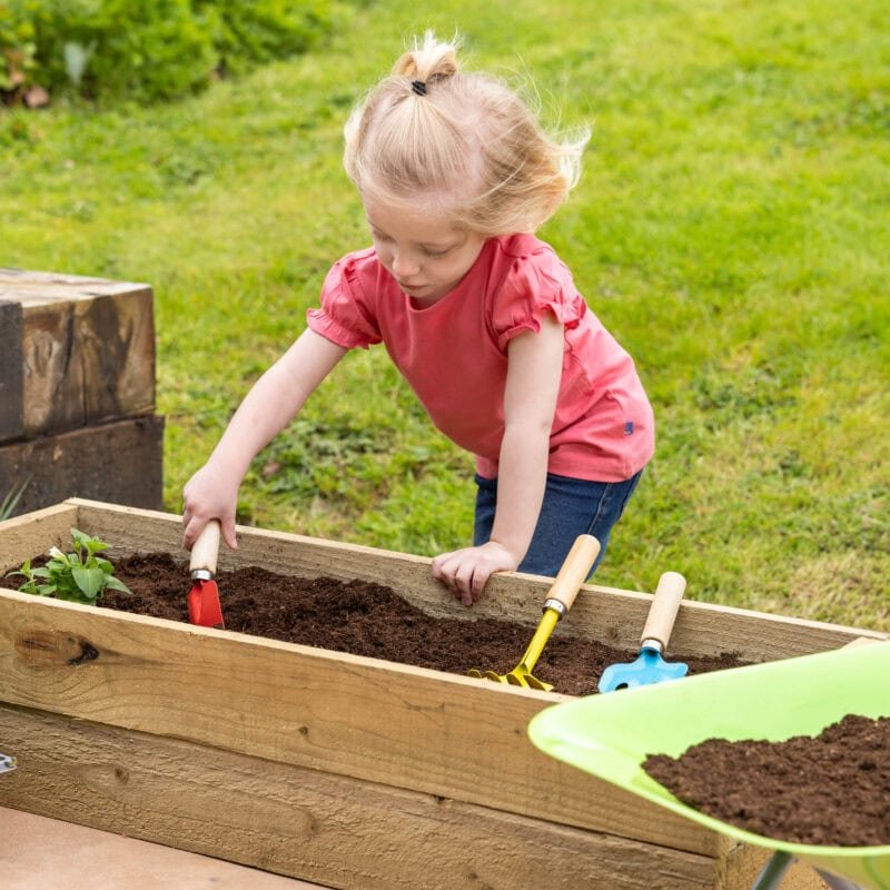 Une petite fille plante un jardin avec des outils de jardinage colorés dans une boîte à fleurs en bois à l'extérieur.