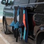 Rugzakken voor buitenactiviteiten hangen aan autodeur, met surfplank klaar voor strandplezier.